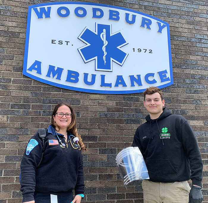 Image of student presenting homemade face shields to ambulance driver in front of Woodbury Amulance sign